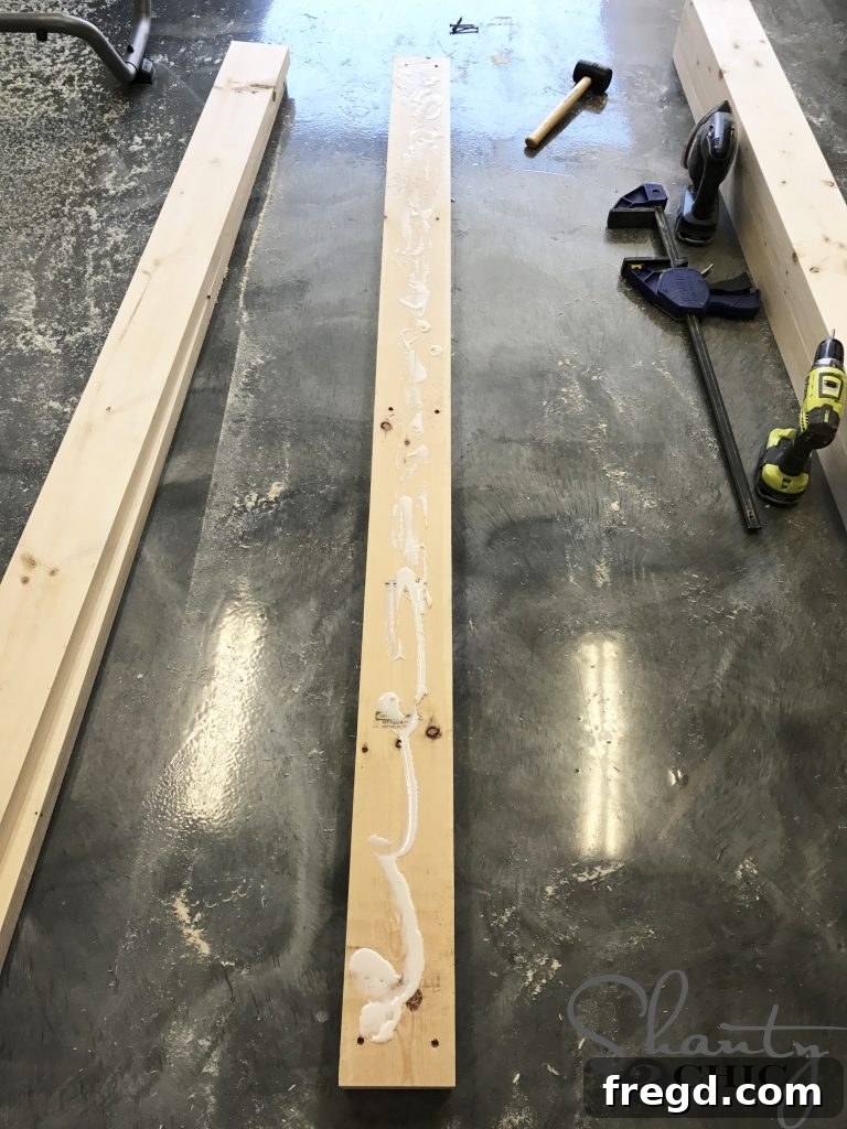 Applying wood glue to the underside of a 2x6 board, preparing it for stacking and assembly to create the side frame of the DIY lounge chair.