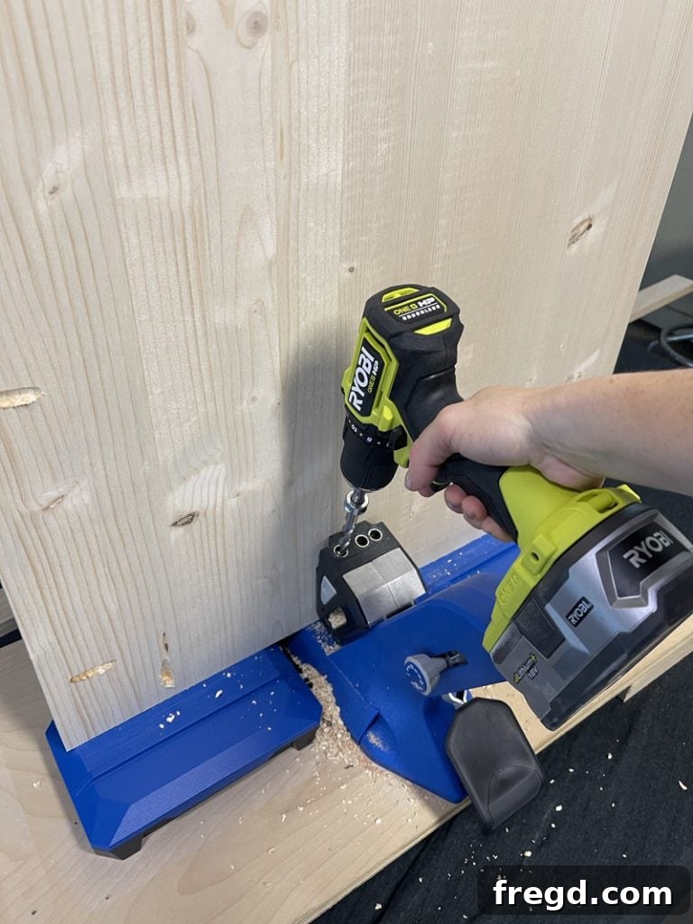 A hand operating a drill with a Kreg bit, creating pocket holes along the edge of a pine project panel.