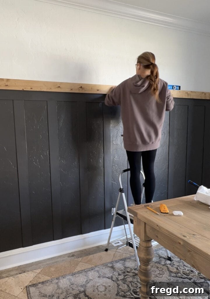 Transform Your Home with DIY Board and Batten Paneling 15 Attaching the stained 1x4 wood trim board to the top of the painted board and batten wall, ensuring it's level and secured into studs.