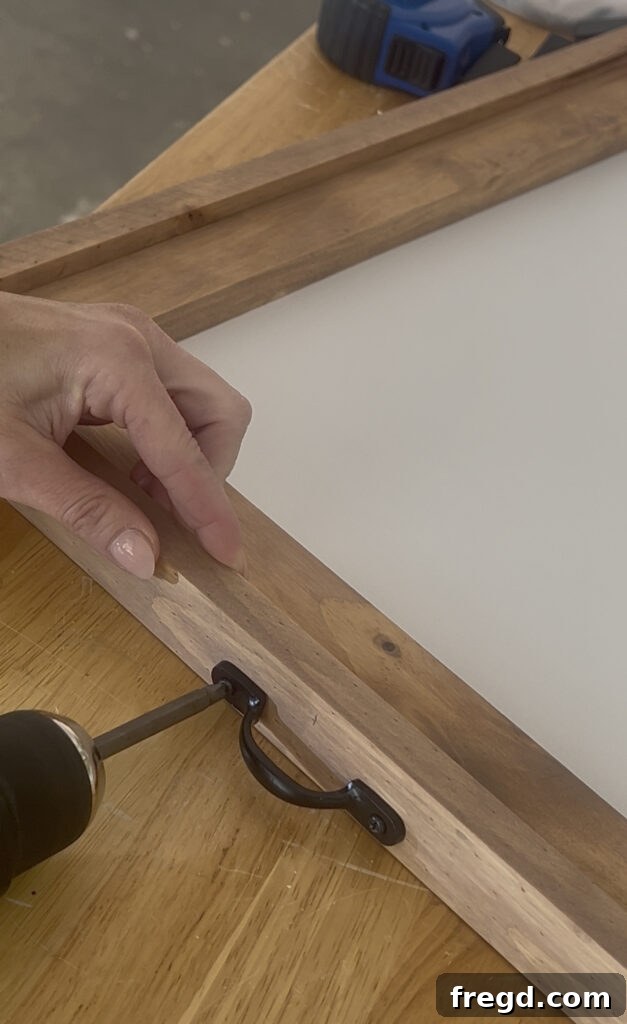 A black metal drawer pull being attached to the top center of a stained wooden frame with wood screws.