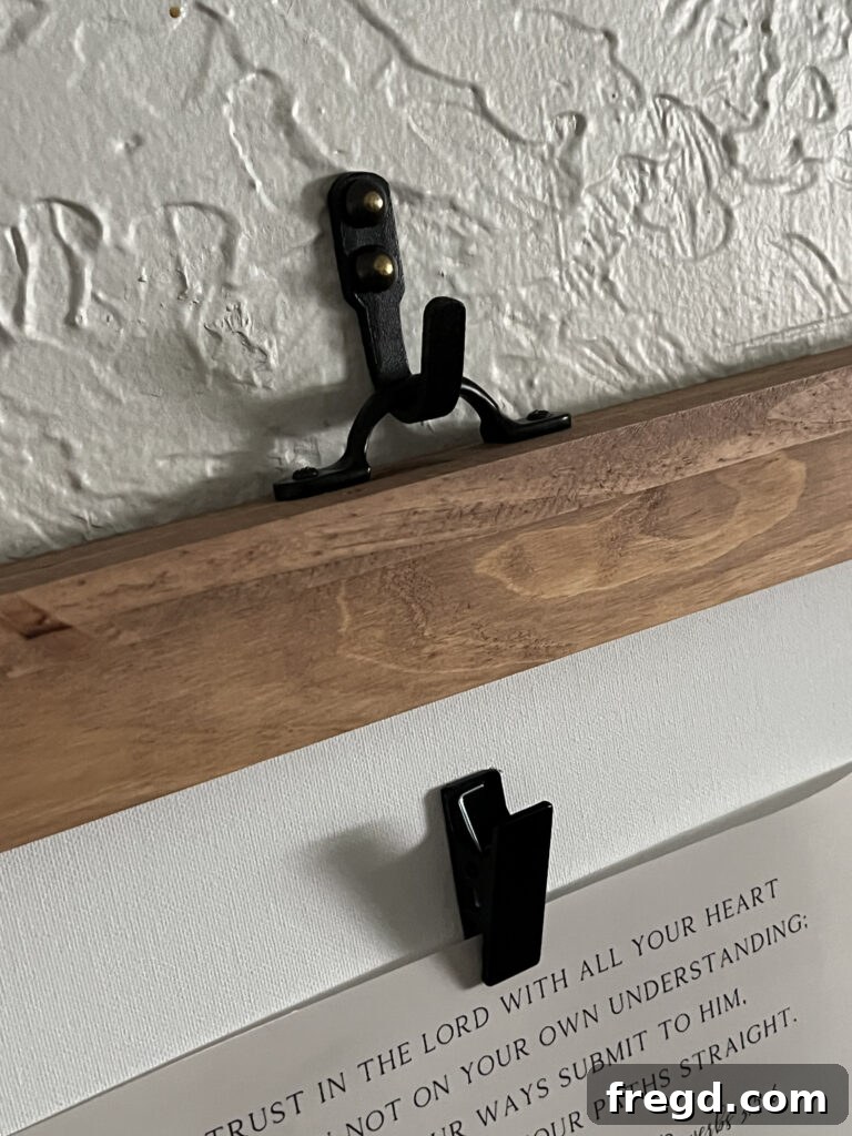 A decorative black metal wall hook inserted through a drawer pull on a wooden frame, and then secured to the wall with a brass thumbtack, creating an illusion of hanging.